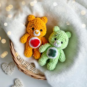 May include: Two handmade crochet teddy bears in a wicker basket. One bear is yellow with a red and white belly, the other is light green with a dark green and white belly. The bears are on a white blanket.