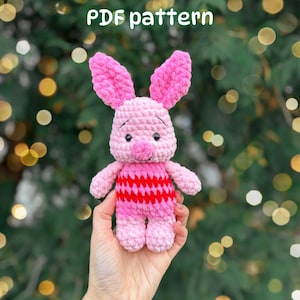 May include: A crochet plush toy of a small pink pig character with long ears, black eyes, and a round snout. The toy features a body with horizontal stripes in light pink and dark red. The text PDF pattern is visible at the top of the image.