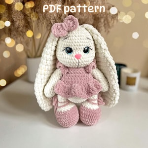 Puede incluir: Una muñeca de conejo tejida a ganchillo con hilo blanco y orejas largas, que lleva un vestido rosa polvoriento, zapatos a juego con lazos y un pequeño moño en la cabeza. La muñeca tiene ojos azules y nariz rosa. El texto PDF pattern es visible.