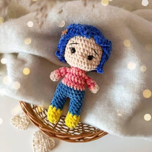 May include: A handmade crochet doll featuring blue hair with a small dragonfly hair clip, a light beige face with black embroidered eyes, a pink long-sleeved shirt, blue pants, and bright yellow boots, crafted from soft textured yarn.