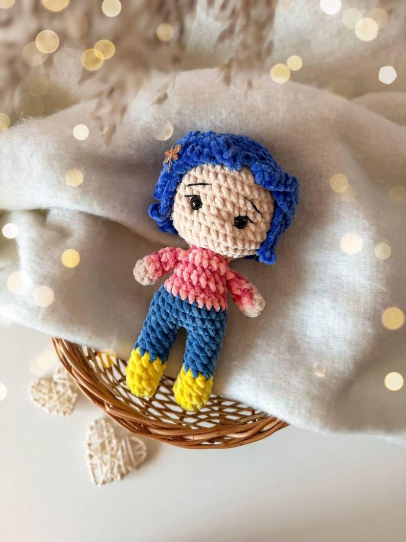 May include: A handmade crochet doll featuring blue hair with a small dragonfly hair clip, a light beige face with black embroidered eyes, a pink long-sleeved shirt, blue pants, and bright yellow boots, crafted from soft textured yarn.