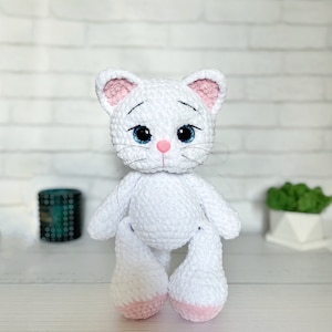Puede incluir: Mu&ntilde;eca de gato de ganchillo blanco hecha a mano con orejas internas rosas, nariz rosa y ojos azules. El gato tiene bigotes negros y est&aacute; sentado erguido. Las patas de la mu&ntilde;eca son blancas con suelas rosas. Fondo de pared de ladrillo blanco.