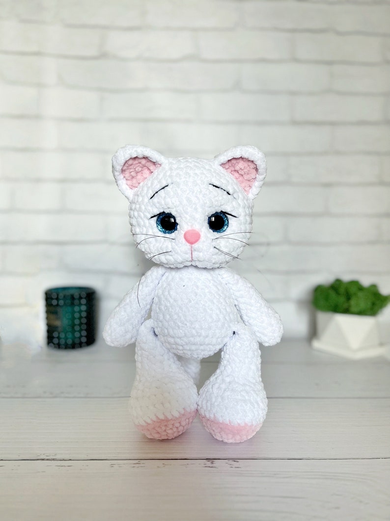 Puede incluir: Mu&ntilde;eca de gato de ganchillo blanco hecha a mano con orejas internas rosas, nariz rosa y ojos azules. El gato tiene bigotes negros y est&aacute; sentado erguido. Las patas de la mu&ntilde;eca son blancas con suelas rosas. Fondo de pared de ladrillo blanco.
