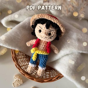 Könnte beinhalten: Gehäkelte Luffy-Puppe mit beigem Gesicht, schwarzen Haaren und einem Strohhut mit rotem Band. Die Puppe trägt eine rote Weste, blaue Jeans und eine gelbe Schärpe. Der Text "PDF PATTERN" steht oben. Die Puppe befindet sich in einem geflochtenen Korb.