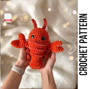 Puede incluir: Un juguete de amigurumi de langosta tejido a crochet con ojos negros y una boca sonriente. La langosta es naranja y está siendo sostenida por las manos de una persona.