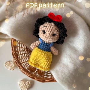 Pode incluir: Uma boneca de crochê com cabelo preto e um laço vermelho, um corpete azul e uma saia amarela. A boneca tem pele clara, traços faciais bordados e sapatos castanhos. O texto PDF pattern é visível no topo da imagem.