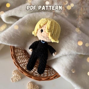 May include: A handmade crochet doll with a blonde head and black outfit, sitting in a small wicker basket. The doll has a light skin tone and is wearing a black suit with a blue tie. The text "PDF PATTERN" is at the top.