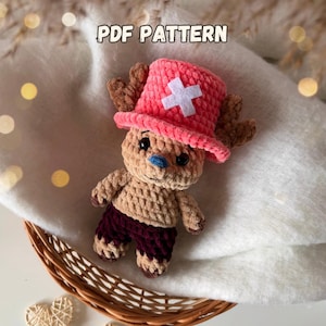May include: A crocheted plush toy of a cartoon character, with a pink hat and white cross, brown antlers, and burgundy shorts. The toy is beige and blue, and the text "PDF PATTERN" is visible.