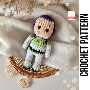 May include: A handmade crochet doll of a cartoon character, featuring a light tan face, purple hair, and a green, white, and black spacesuit. The doll is displayed in a small woven basket on a white surface. The text "CROCHET PATTERN" is visible.