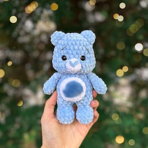 May include: A light blue crochet teddy bear with a white heart-shaped nose and a blue belly. The plush toy is held in a hand against a blurred background of green and gold bokeh lights. The bear has black eyes and a soft, textured appearance.