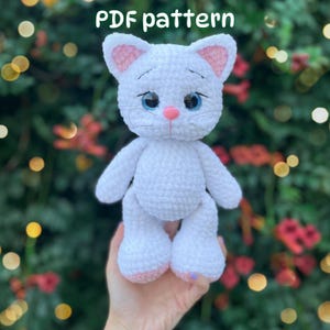 May include: A white crocheted cat plush toy with large blue eyes, a pink nose, and pink inner ears. The text 'PDF pattern' is visible at the top. The cat has embroidered whiskers and a soft, textured yarn finish, standing in a vertical position.