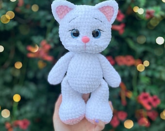 May include: A white crocheted cat plush toy with large blue eyes, a pink nose, and pink inner ears. The text 'PDF pattern' is visible at the top. The cat has embroidered whiskers and a soft, textured yarn finish, standing in a vertical position.