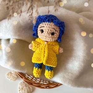May include: A handmade crochet doll featuring a beige yarn head with dark blue hair, wearing a bright yellow textured coat over blue pants and yellow boots. The doll has embroidered black eyes and eyebrows, with a small white button detail on the coat.