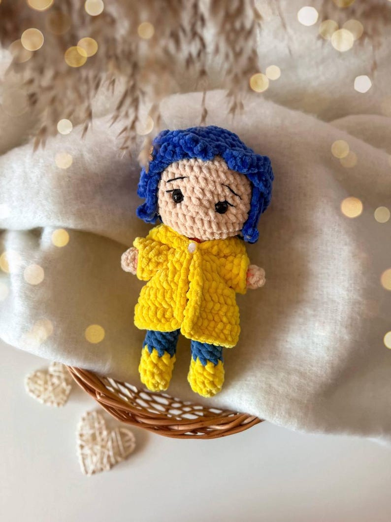 May include: A handmade crochet doll featuring a beige yarn head with dark blue hair, wearing a bright yellow textured coat over blue pants and yellow boots. The doll has embroidered black eyes and eyebrows, with a small white button detail on the coat.