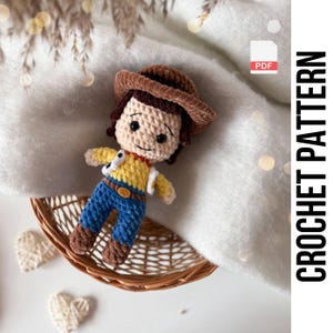 May include: A crocheted doll of a cowboy character, with a brown hat, yellow shirt, blue trousers, and a brown waistcoat. The doll is in a small woven basket. The image also includes the text "CROCHET PATTERN".