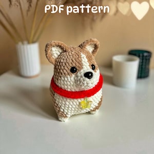 Può includere: Un peluche corgi all'uncinetto realizzato con filato marrone e bianco, con occhi in plastica nera e naso nero. Il corgi indossa un collare rosso brillante con un piccolo ciondolo a forma di stella gialla traslucida al centro. Il testo recita PDF pattern.