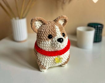May include: A crochet corgi plush toy made with brown and white yarn, featuring black plastic eyes and a black nose. The corgi wears a bright red collar with a small, translucent yellow star-shaped charm hanging from the center. Text reads PDF pattern.