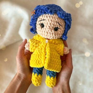 May include: A handmade crochet doll featuring a character with textured blue yarn hair, a small orange dragonfly hair clip, and black safety eyes. The doll wears a bright yellow hooded raincoat with a single white button and blue and yellow boots.