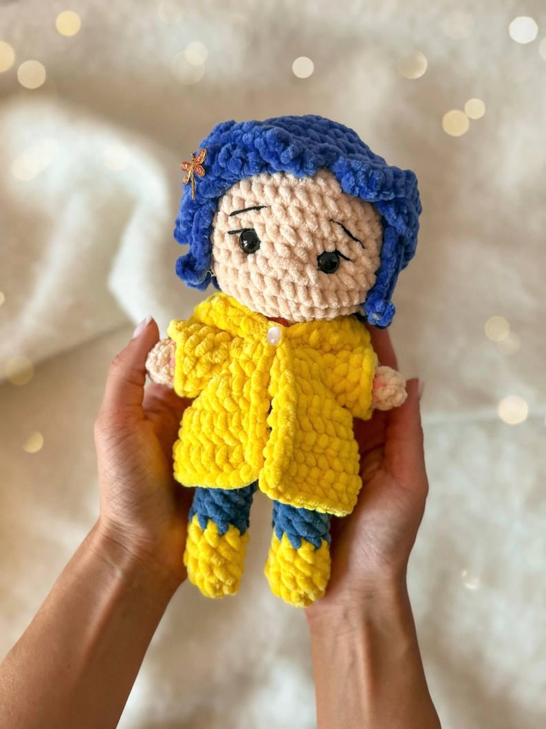 May include: A handmade crochet doll featuring a character with textured blue yarn hair, a small orange dragonfly hair clip, and black safety eyes. The doll wears a bright yellow hooded raincoat with a single white button and blue and yellow boots.