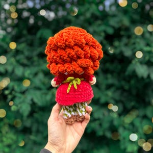 May include: A crocheted doll with voluminous, textured orange yarn hair, wearing a red dress with a small green bow tied at the waist. The doll has patterned white and green legs and brown feet, crafted from soft yarn in a handmade style.