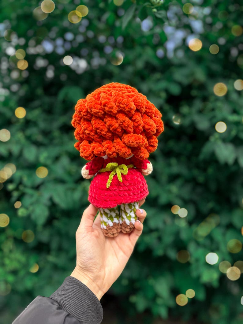 May include: A crocheted doll with voluminous, textured orange yarn hair, wearing a red dress with a small green bow tied at the waist. The doll has patterned white and green legs and brown feet, crafted from soft yarn in a handmade style.
