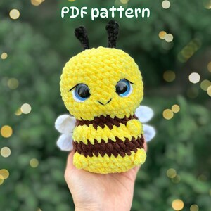 May include: A crochet bee plush toy featuring a yellow and brown striped body, black antennae, large blue eyes, and small white wings. The text PDF pattern is displayed at the top of the image. The plush has a soft, textured yarn finish.