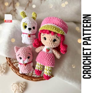 May include: A collection of handmade crochet dolls and animals. The set includes a pink doll with red hair, a white dog with yellow ears, and a pink cat. The doll is wearing a pink dress and a hat. The text "CROCHET PATTERN" is visible.