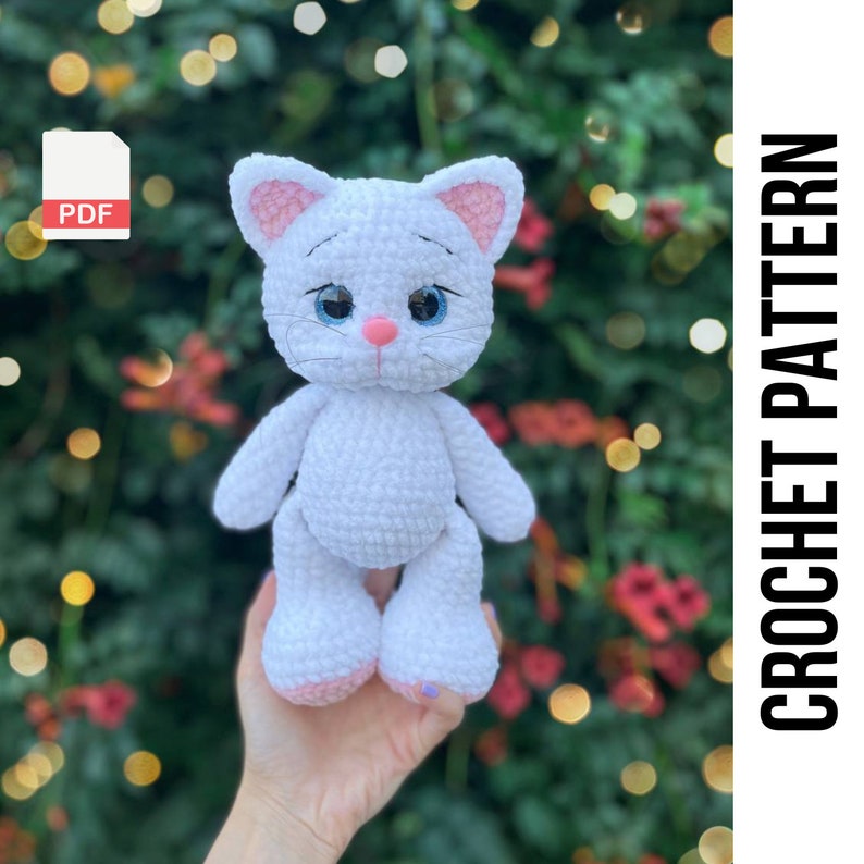 Puede incluir: Una mu&ntilde;eca de gato de ganchillo blanco con el interior de las orejas, la nariz y las almohadillas rosas. El gato tiene ojos azules y una expresi&oacute;n dulce. El texto "CROCHET PATTERN" est&aacute; incluido.