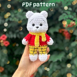 Könnte beinhalten: Ein weißes gehäkeltes Bären-Stofftier mit schwarzen Augen und Nase, das einen roten Pullover, einen gelben Schal und eine gelb karierte Hose trägt. Der Text "PDF pattern" ist oben im Bild sichtbar. Der Bär hat weiße Pfoten und Ohren.