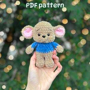 Puede incluir: Un peluche de ratón tejido a ganchillo con pelaje color canela, orejas grandes con detalles internos rosas, ojos negros y una pequeña nariz negra. El ratón lleva un suéter azul. El texto PDF pattern es visible en la parte superior.