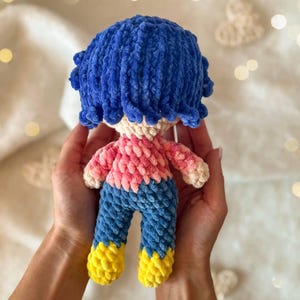 May include: A small, handmade crochet doll featuring a textured blue yarn hairstyle, a light beige head, a pink and white variegated yarn shirt, blue yarn pants, and bright yellow yarn shoes, crafted with a soft, plush, and fuzzy yarn texture.