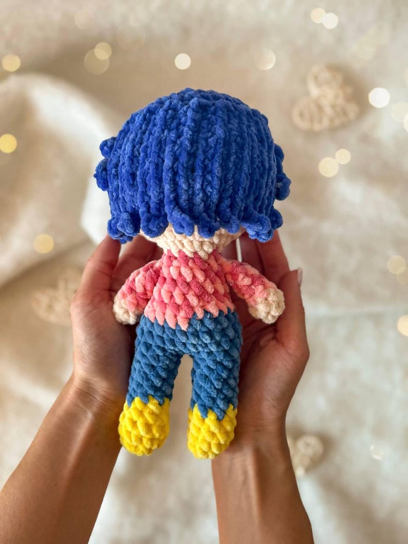 May include: A small, handmade crochet doll featuring a textured blue yarn hairstyle, a light beige head, a pink and white variegated yarn shirt, blue yarn pants, and bright yellow yarn shoes, crafted with a soft, plush, and fuzzy yarn texture.