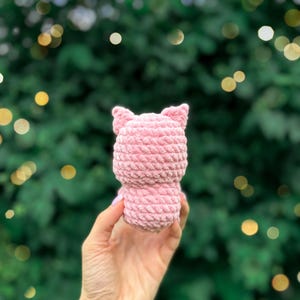 May include: A small, light pink plush toy crafted from thick, textured yarn. The object features a rounded body and two pointed ears at the top, showcasing a consistent crocheted stitch pattern throughout its entire surface.