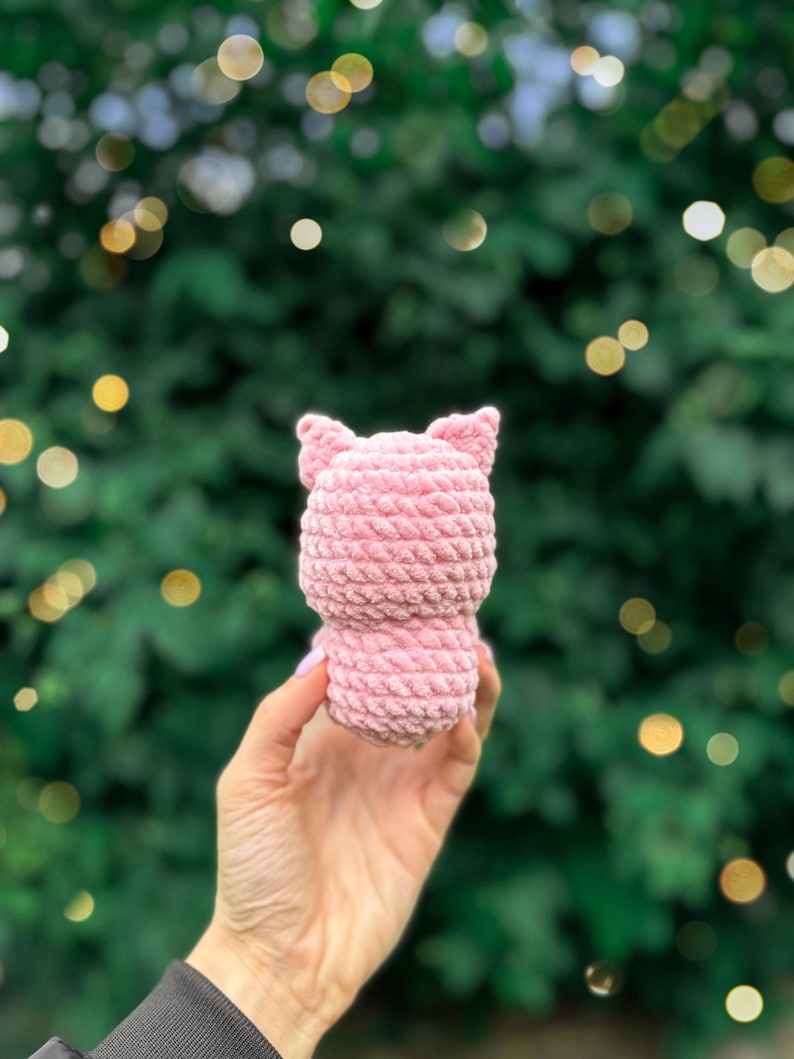 May include: A small, light pink plush toy crafted from thick, textured yarn. The object features a rounded body and two pointed ears at the top, showcasing a consistent crocheted stitch pattern throughout its entire surface.