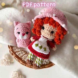 May include: A crochet doll with bright orange curly hair, wearing a pink hat with white accents and a white apron over a red dress, accompanied by a small pink crochet cat. The text PDF pattern is visible at the top of the image.