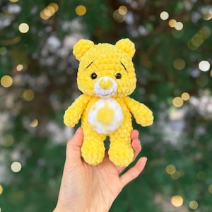 May include: A handmade yellow plush bear with a white belly and a smiling face. The bear is made of soft, textured yarn and has small black eyes. The bear is being held in a person's hand.