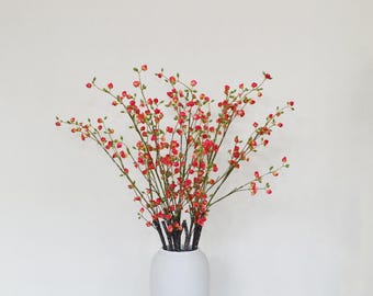 May include: A bouquet of artificial branches featuring numerous small, vibrant red flower buds and green leaves, arranged in a simple white vase. The thin, dark brown stems extend outward in a fan shape, showcasing a dense cluster of blossoms.