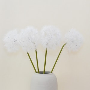 May include: Four white dandelion flower stems in a grey ceramic vase against a white background.