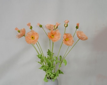 May include: A bouquet of peach-colored artificial poppy flowers with green leaves in a gray vase.