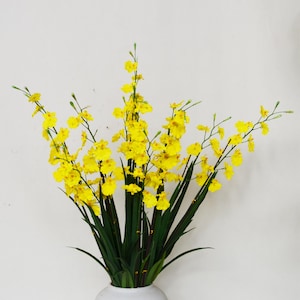 Puede incluir: Un arreglo de orquídeas amarillas con hojas verdes en un jarrón blanco. Las flores amarillas brillantes crean un ambiente alegre. Ideal para la decoración del hogar o como regalo.
