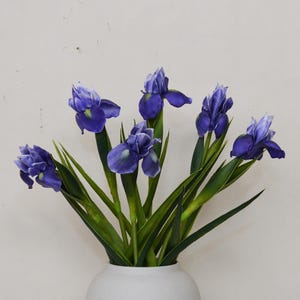 May include: A bouquet of artificial irises in a white vase. The flowers are a vibrant purple with green stems and leaves. The vase is a simple, rounded shape, and the background is a plain white wall.