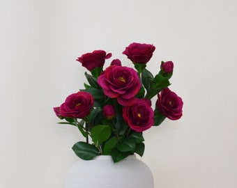 May include: A bouquet of deep pink artificial flowers in a white ceramic vase. The flowers have a ruffled appearance and are arranged in a cluster.