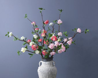 May include: A floral arrangement featuring branches with pink, red, and white blossoms and green leaves. The branches are in a textured, off-white vase with dark accents. The background is a solid, muted blue.