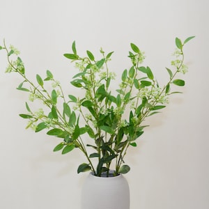 Puede incluir: Un ramo de ramas verdes artificiales con pequeñas flores blancas en un jarrón de cerámica blanco.