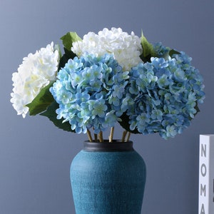 May include: A bouquet of blue and white hydrangeas in a teal ceramic vase. The vase is sitting on a table with two books.