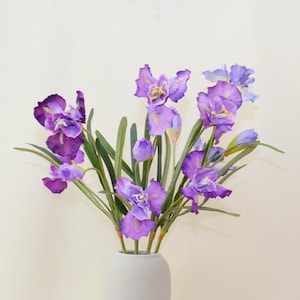 May include: A bouquet of purple iris flowers in a white vase. The flowers have delicate petals and are arranged in a natural, flowing style.