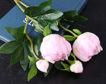 以下が含まれることがあります： 緑の葉が付いた人工のピンクの牡丹の花束。花は花瓶に活けられており、家やオフィスに飾る準備ができています。