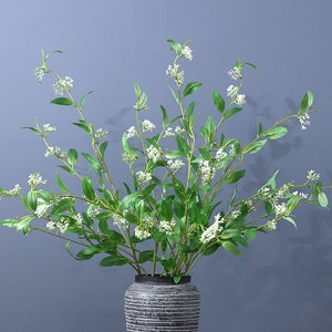 Puede incluir: Un ramo de flores blancas con hojas verdes dispuestas en un jarrón de cerámica gris. Las flores son delicadas y tienen una textura suave y plumosa.