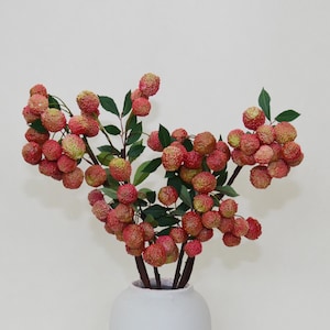 May include: A bouquet of artificial lychee branches with red and green leaves in a white vase.