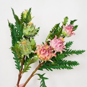 Op de afbeelding: Een boeket kunstbloemen met groene en roze bloemen en gele centra. De bloemen hebben een unieke, getextureerde uitstraling en zijn geplaatst tegen groene, varenachtige bladeren. De stelen zijn bruin.